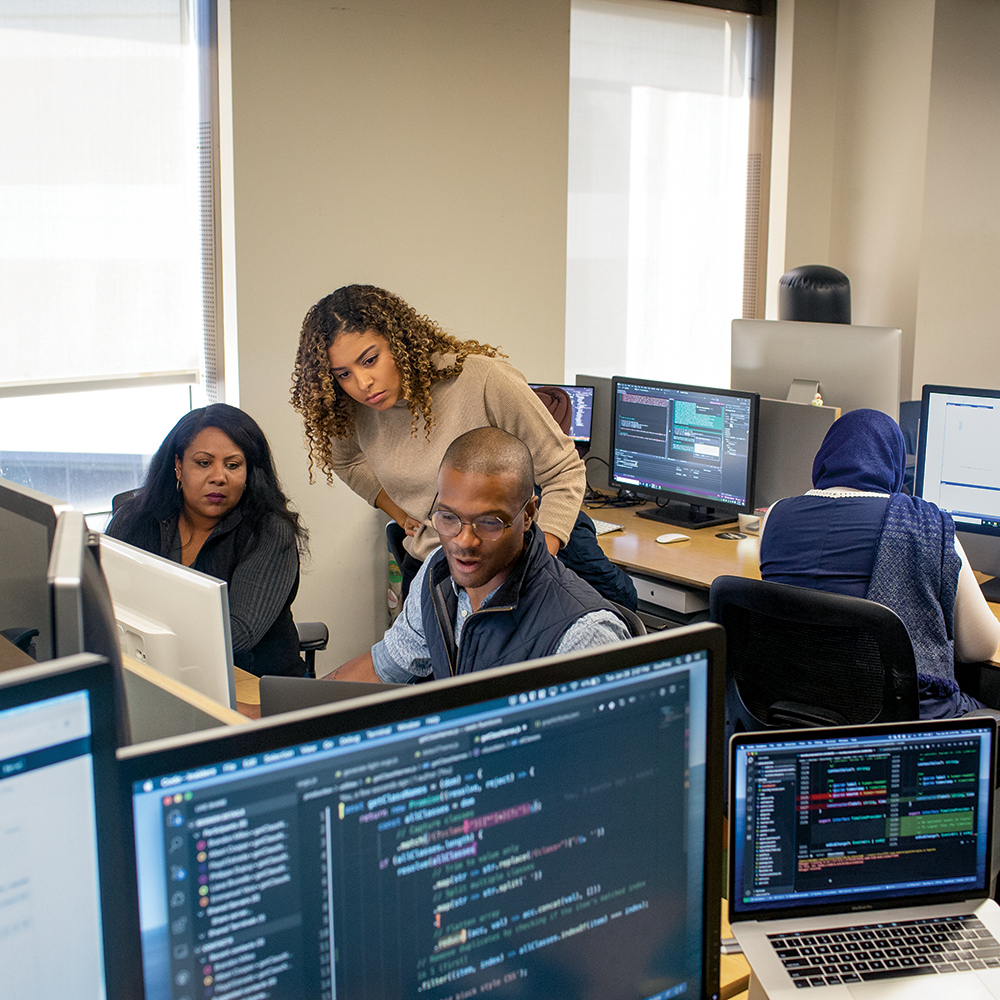 Three people gathered around a computer in an office space filled with multiple monitors. Three people gathered around a computer in an office space filled with multiple monitors.