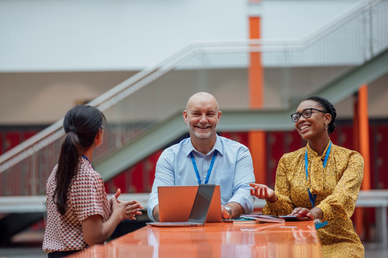 Three educators with laptops sit at a table and collaborate in a school common area.