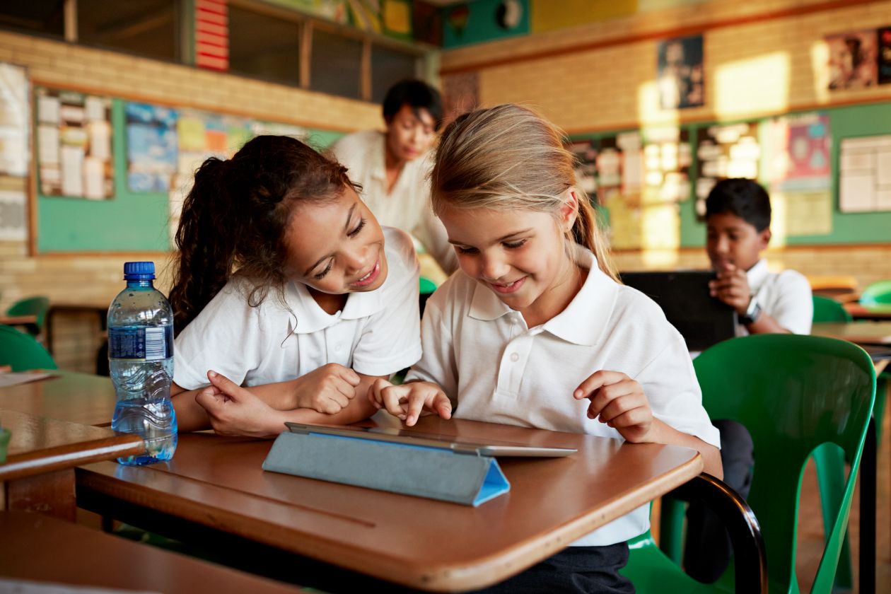 Two young students playing on a tablet in a school classroom.