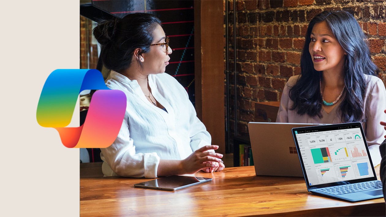 Two people sitting at a table looking at a laptop. Microsoft Copilot logo included.